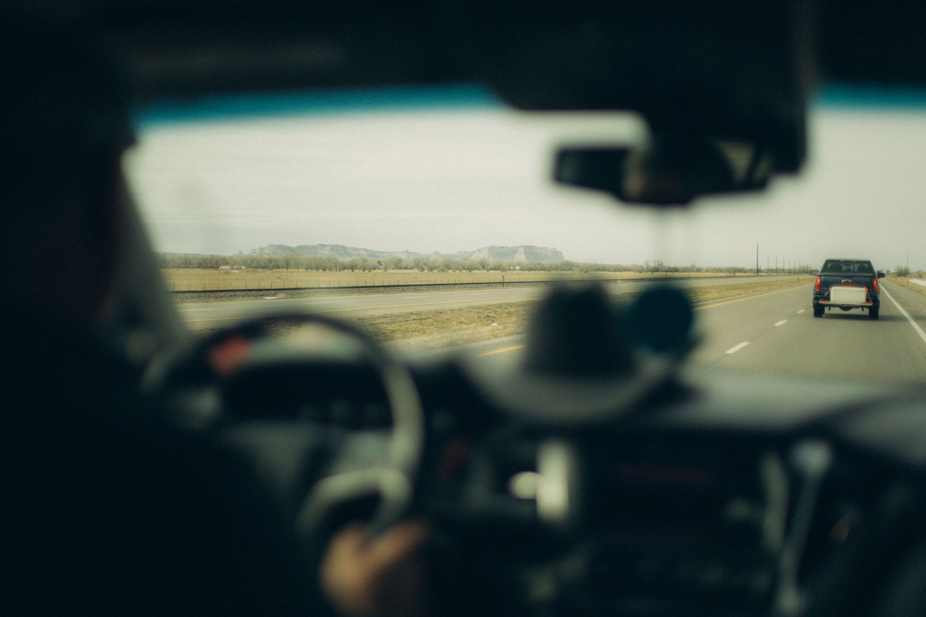 View from inside a car driving on a highway with another vehicle ahead and flat desert landscape with mountains in the distance